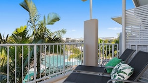 A balcony with a view of a swimming pool, palm trees, and a building in the background. - Resort & Spa 6316 withTropical Pool Views - 230m to Beach (KINGSCLIFF)