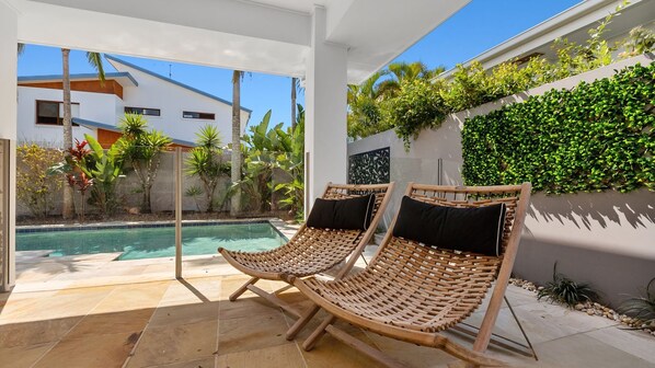 A poolside area with two wicker lounge chairs, a tiled patio, and a view of a modern building and lush greenery.