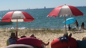 En strand med folk som kopplar av under parasoller, en sandstrand och segelbåtar på vattnet.