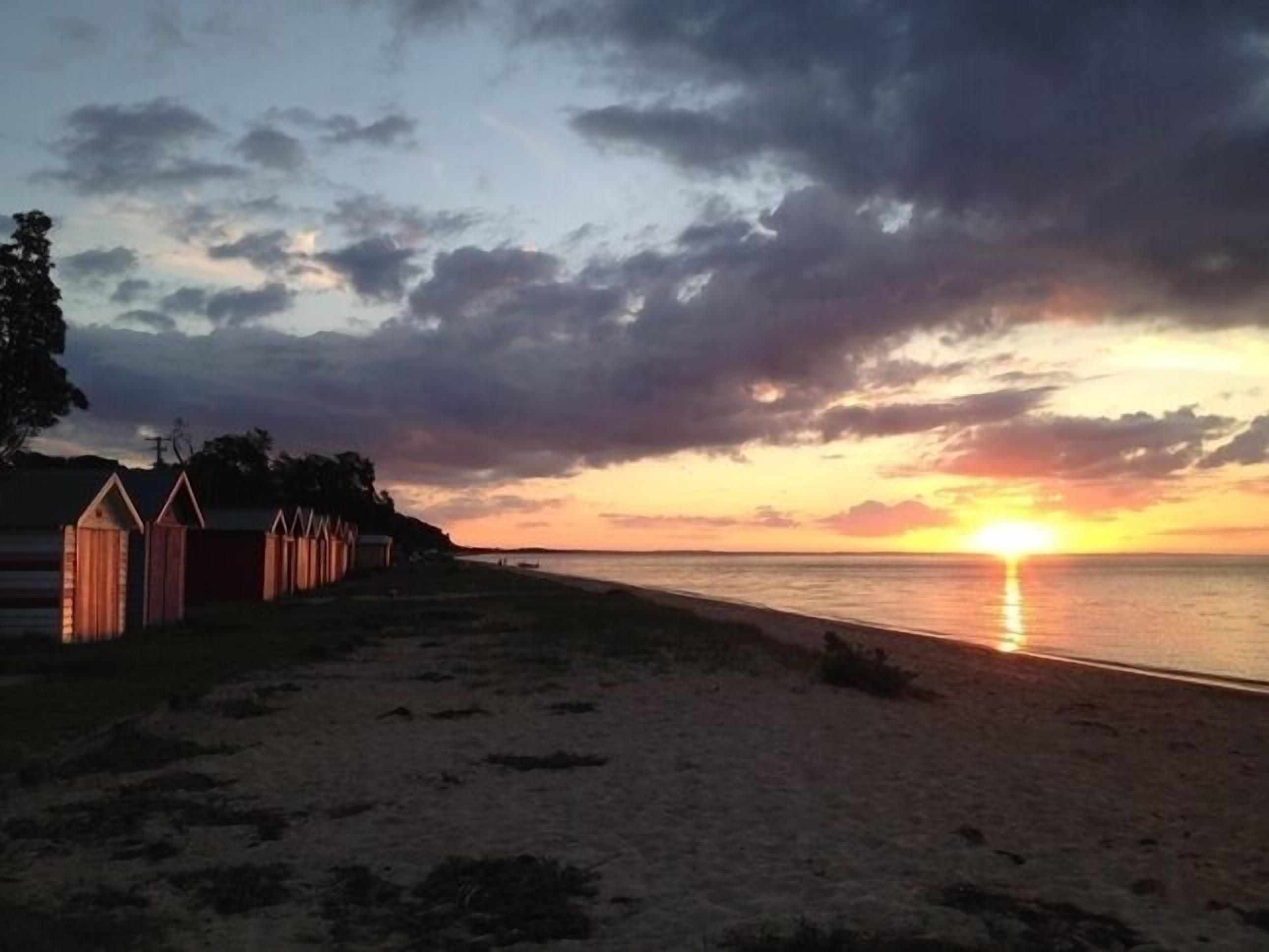 Ein Strand mit Holzgebäuden, vermutlich Strandkörben, und einem Sonnenuntergang über dem Wasser.