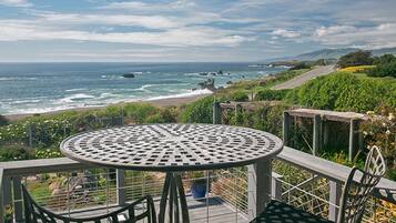 A round metal table with chairs on an outdoor deck, overlooking a beach and ocean.