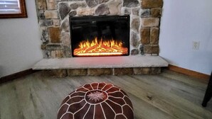 Ein Wohnzimmer mit Kamin aus Stein, einem flachbildfernseher darüber und einem dekorativen Hocker davor.
