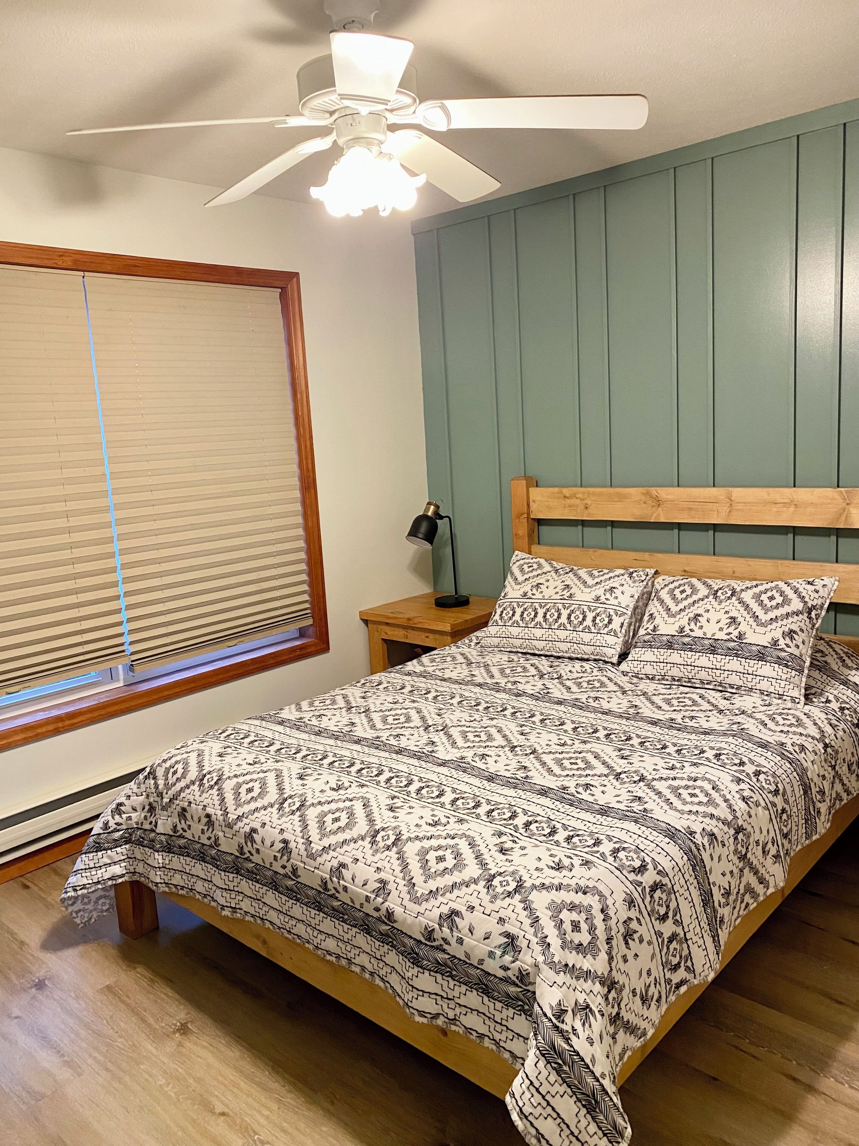 Un dormitorio con cama de madera, ropa de cama estampada, ventilador de techo, mesita de noche con lámpara y ventana con persianas.