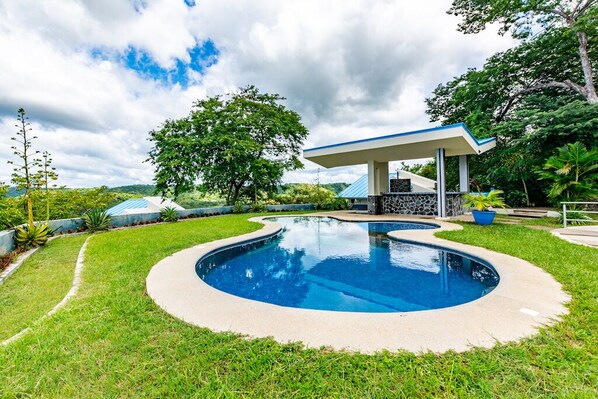 A swimming pool with a curved edge, surrounded by a well-maintained lawn and a gazebo-like structure.