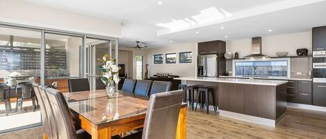 A modern kitchen with a central island, a dining area with a glass table, and a view through a glass door.