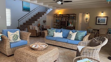 Un salon spacieux avec un canapé bleu, des meubles en osier, un escalier et un ventilateur de plafond.