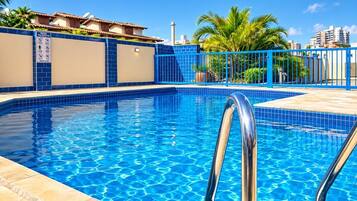 Piscina azul límpida com escada de metal, cercada por uma grade azul e palmeiras, com edifícios ao fundo.