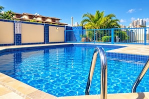 Una piscina azul cristalina con una escalera de metal, rodeada por una cerca azul y palmeras, con edificios al fondo.