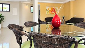 Un coin repas avec une table ronde en verre entourée de chaises en osier, un vase rouge servant de centre de table et un mur jaune orné d’un tableau floral.
