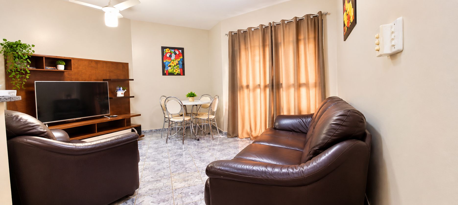 Uma sala de estar com televisão, sofás de couro, mesa de jantar com cadeiras e janela com cortinas.