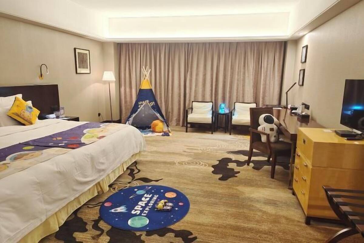 A hotel room with a bed, a desk, a chair, a dresser, a lamp, a television, a space-themed rug, and a teepee decoration.