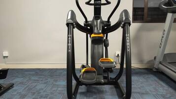 An exercise machine in a room with a blue carpet, a window, and a treadmill in the background.