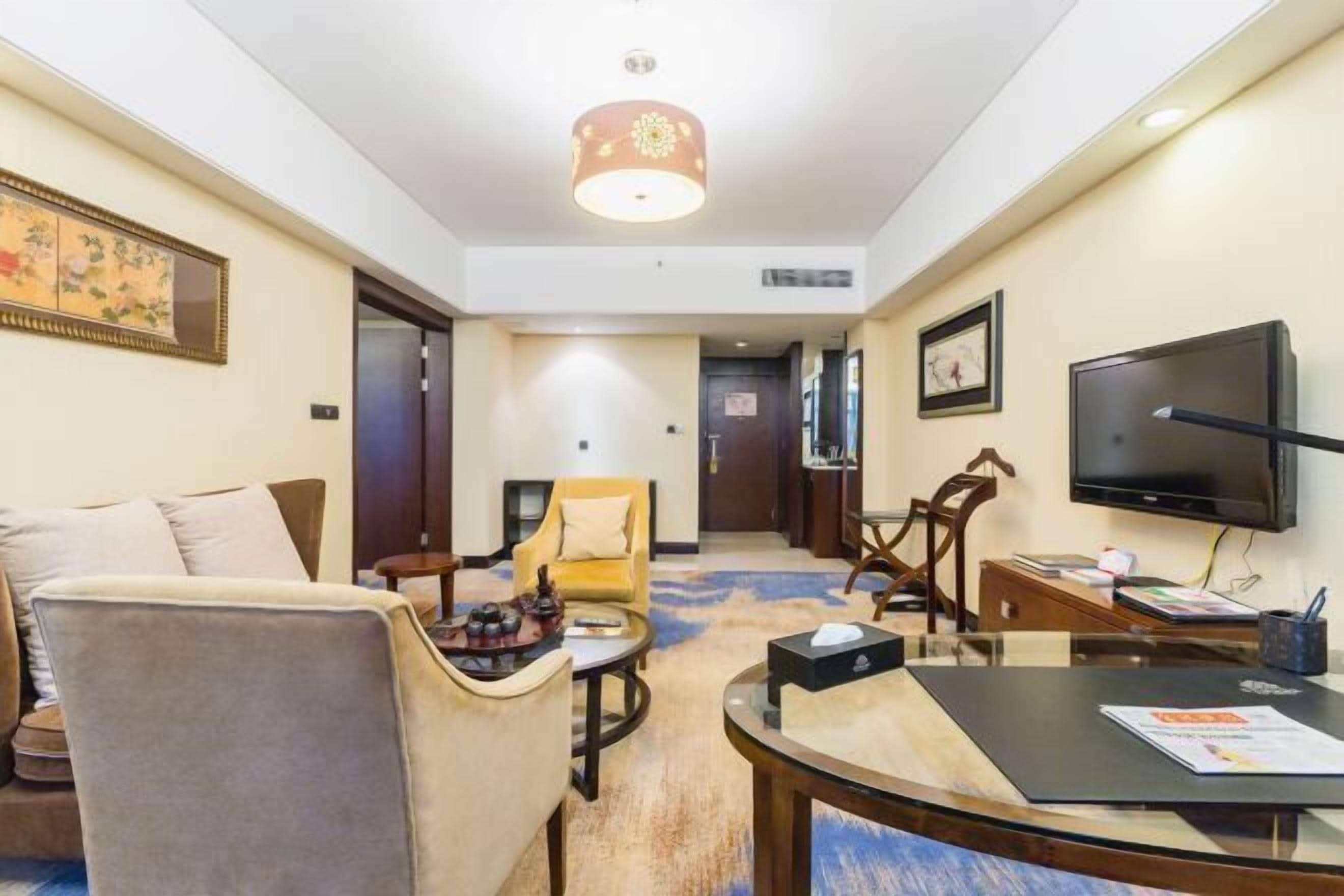 Quarto de hotel com sofá, poltronas, uma mesa pequena, televisão e piso coberto por carpete.