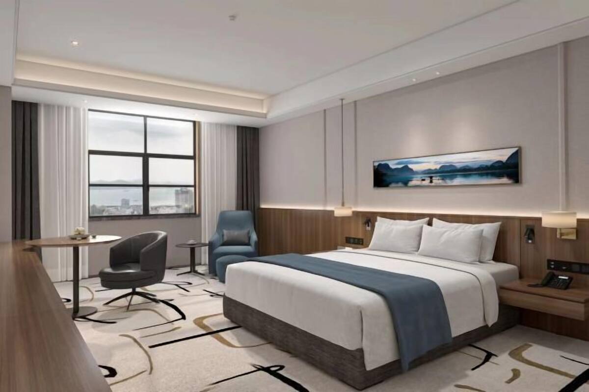 A modern hotel room with a large bed, a desk, a chair, and a view of the city.