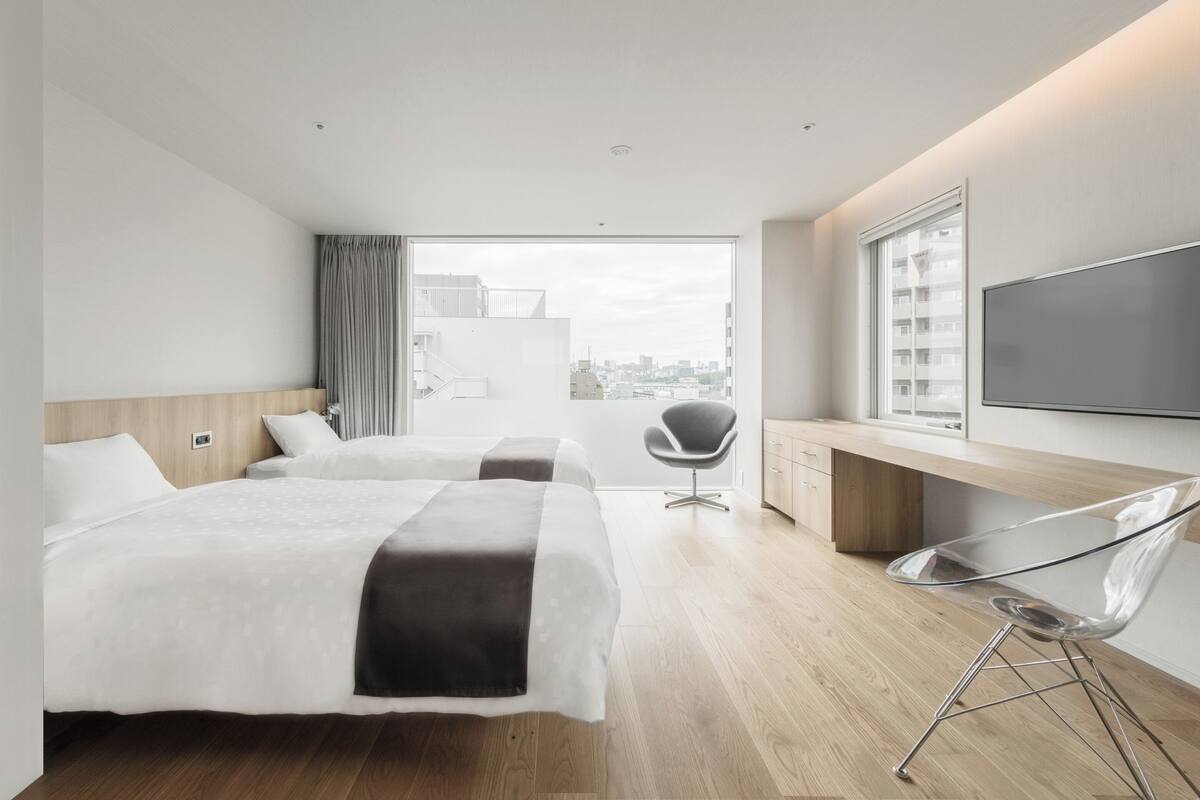 A modern hotel room with two beds, a desk, a chair, and a large window offering a city view.