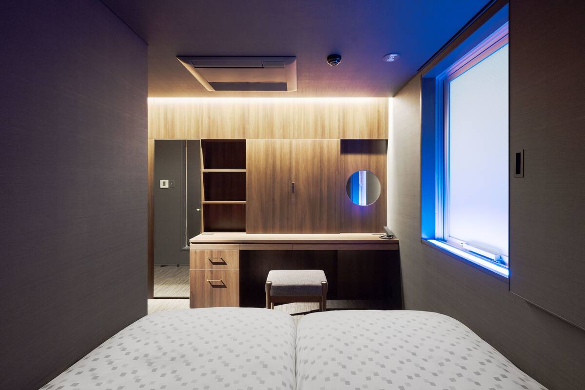 A modern bedroom with a wooden desk, a small stool, a bed with patterned bedding, and a window with blue light.