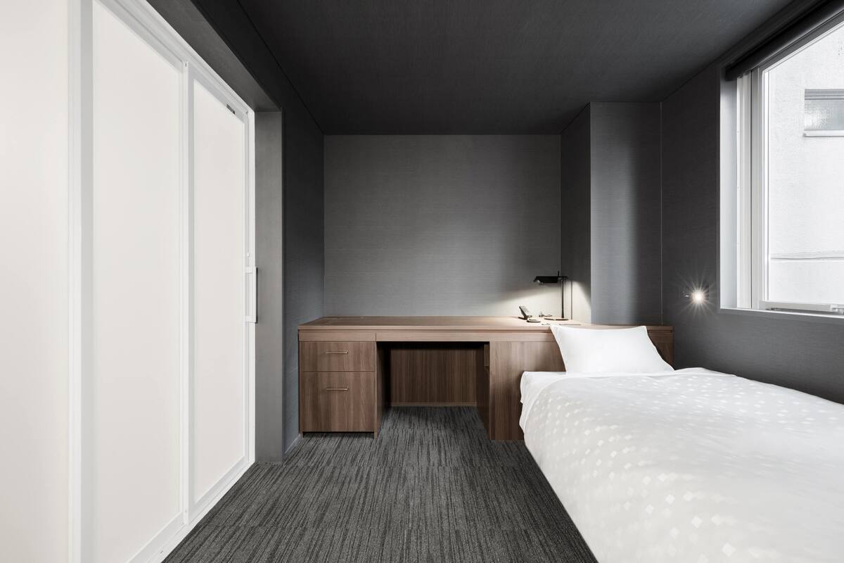 A compact hotel room with a single bed, a desk, and a window.