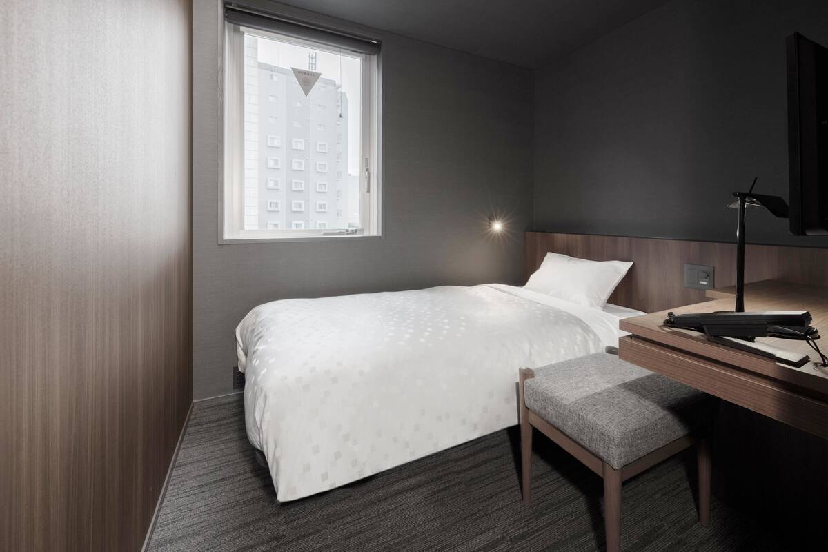 A hotel room with a bed, a desk, a chair, a window with a view of buildings, and a wall-mounted lamp.