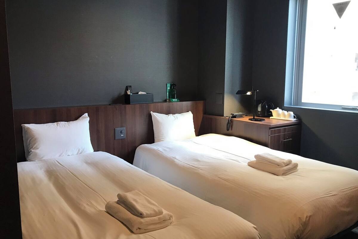 A hotel room with two single beds, a nightstand, a lamp, and a window with a view.