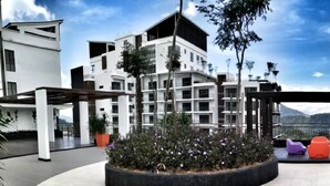 A modern building with a courtyard featuring a planter with plants and flowers, seating areas, and a view of the surrounding landscape. - Midhills at Genting by Self Check In (Genting Highlands)