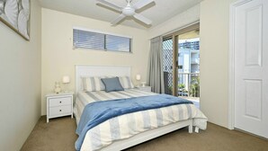 Une chambre à coucher comprenant un lit, des tables de chevet, un ventilateur de plafond, une fenêtre avec des stores et une vue sur un bâtiment à l’extérieur.