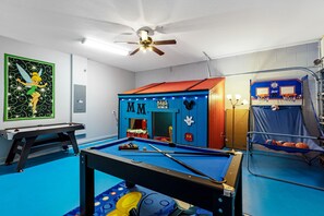 Une table de billard aux motifs colorés, un ventilateur de plafond et un panier de basket dans une pièce dont le sol est bleu et les murs blancs.