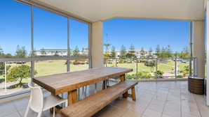 Een balkon met een houten tafel en bank, grote ramen en uitzicht op een golfbaan.