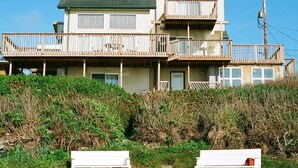 A two-story beachfront house with a balcony, a deck, and a sandy foreground with two white benches. - Shearwater Beach Front Vacation Rental 3000 sqft (Depoe Bay)