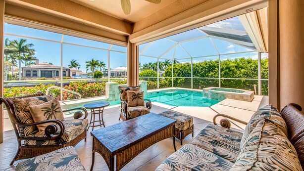 A sunlit room with a pool, wicker furniture, and large windows offering a view of palm trees and a distant building. - Beautifully Furnished Tommy Bahama Inspired Home (Marco Island)