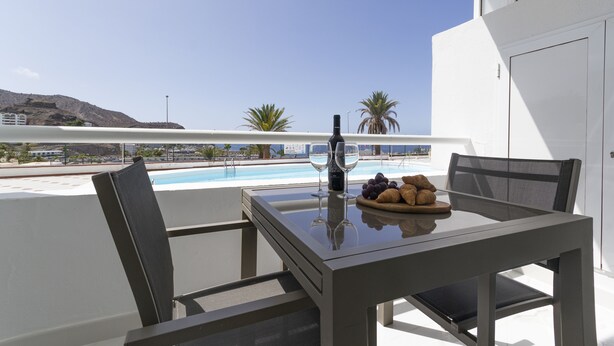 A balcony with a table set for two, overlooking a pool and a mountainous landscape. - Puerto Rico Gran Canaria: Beach and enjoyment at your fingertips (Mogán)