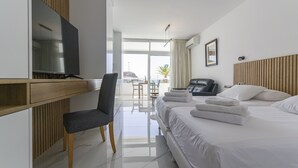 A modern hotel room with a large bed, a desk with a flat-screen TV, a chair, and a view of the outdoors through large windows. - Puerto Rico Gran Canaria: Beach and enjoyment at your fingertips (Mogán)