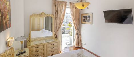 Une chambre d’hôtel avec un lit, une commode, un miroir, une lampe, une télévision et une fenêtre avec des rideaux.