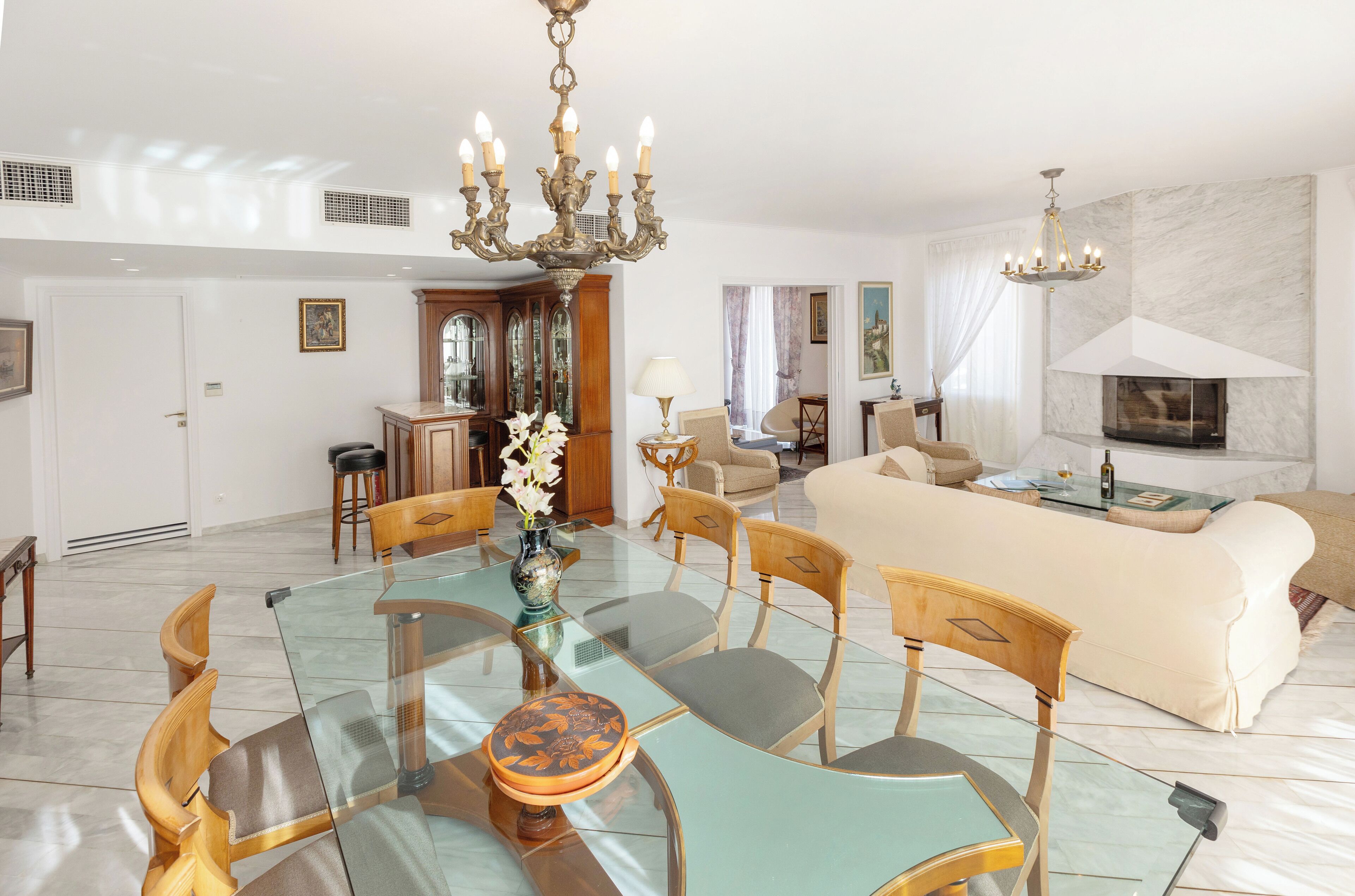 Un salon spacieux avec un grand canapé, un coin repas doté d’une table et de chaises en verre, un lustre et une cheminée.