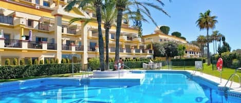 A hotel with a swimming pool, palm trees, and a clear blue sky.