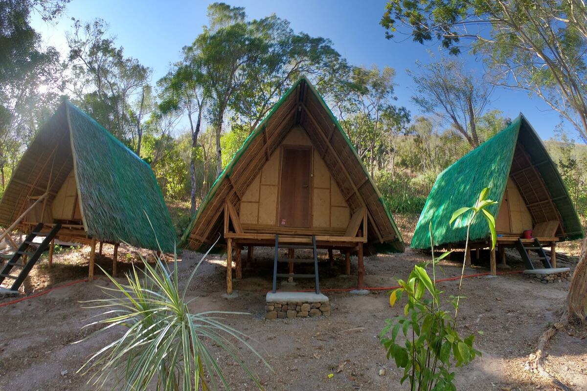 Three thatched-roof huts with wooden frames, surrounded by trees and a dirt ground.