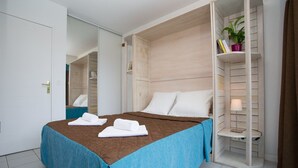 A hotel room with a bed, a nightstand, a shelf with books and a plant, and a door.