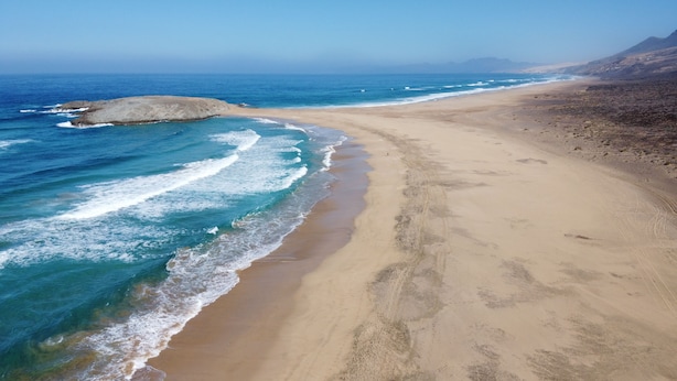 Een strand met helderblauw water, een zandstrand en rotsformaties.