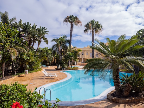A pool area with lounge chairs, palm trees, and a building in the background. - 3 bedroom villa in family complex with pools (Corralejo)