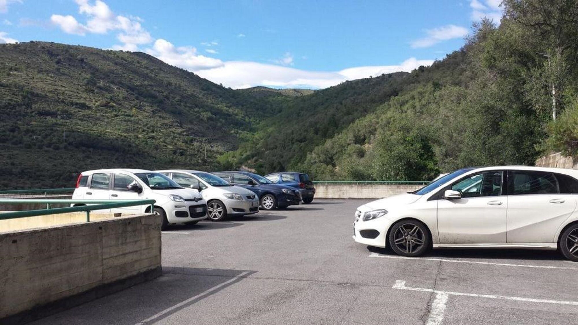 A parking area with cars parked along a green railing, surrounded by lush green hills.