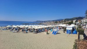 Ein Strand mit vielen Sonnenschirmen und Liegestühlen, einem Spielplatz und Blick auf das Meer und ferne Hügel.