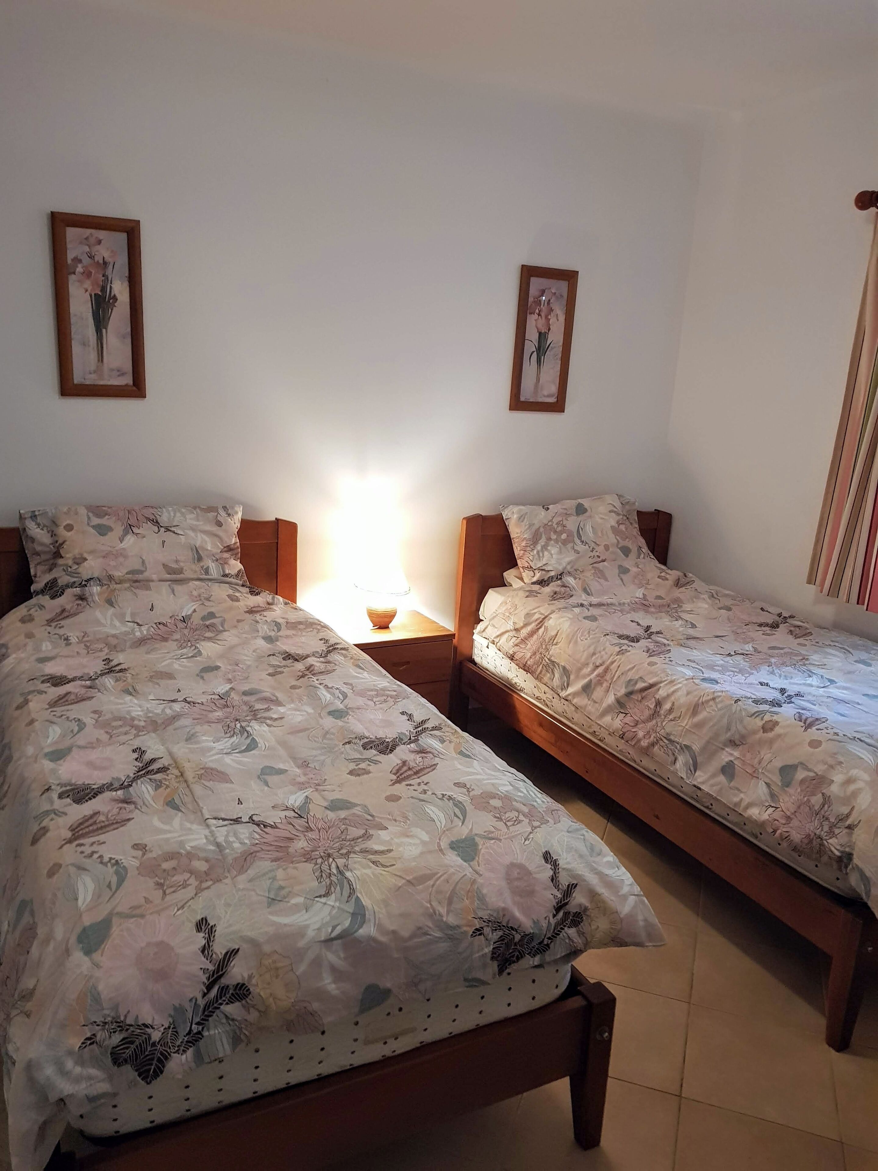 A bedroom with two single beds, each with a floral-patterned bedspread, wooden headboards, and a nightstand with a lamp.