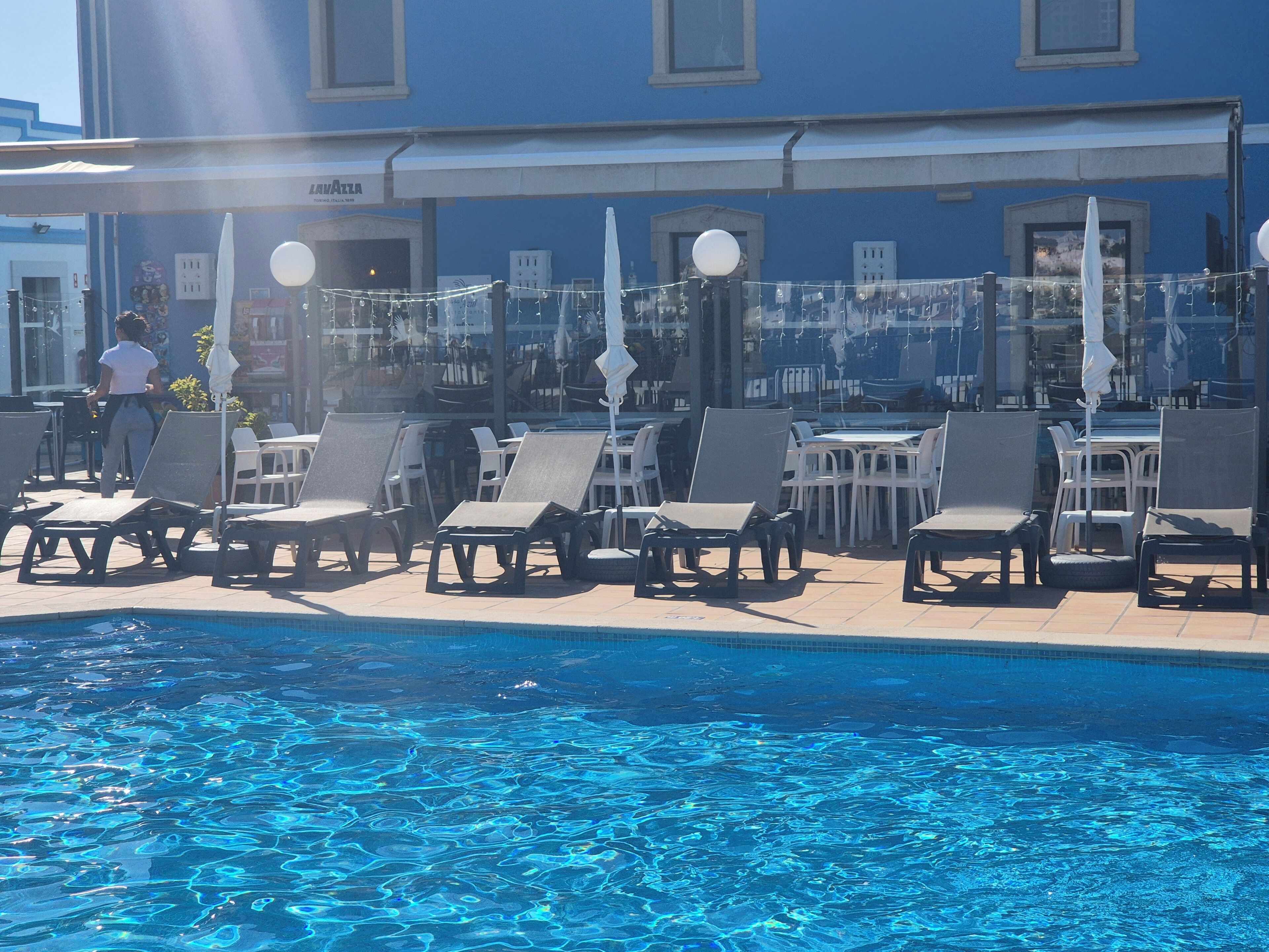 A poolside area with lounge chairs and umbrellas, a clear blue pool, and a building with a blue facade in the background.