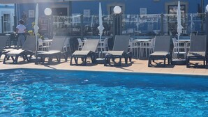 Un espace aménagé au bord de la piscine, avec des chaises longues et des parasols, une piscine à l’eau d’un bleu limpide, et un bâtiment à la façade bleue en arrière-plan.