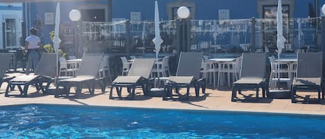 Ett område vid poolen med solstolar och parasoller, en klarblå pool och en byggnad med en blå fasad i bakgrunden.