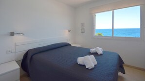 Um quarto com uma cama, duas toalhas brancas, uma mesa de cabeceira, uma janela com vista para o mar e um abajur fixado na parede.
