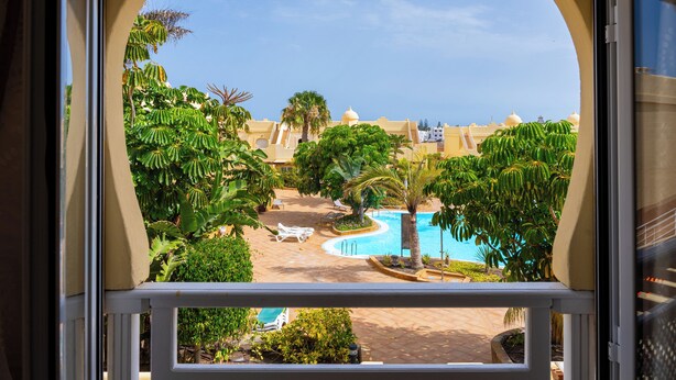 A view of a pool area with lounge chairs, palm trees, and a clear blue sky. - 3 bedroom villa in quiet complex with swimming pools / private wifi / UKTV (Corralejo)