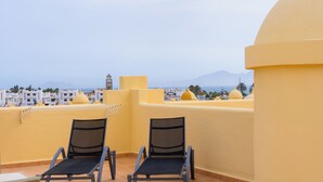 A rooftop terrace with two lounge chairs, a yellow wall, and a view of buildings and mountains in the distance. - 3 bedroom villa in quiet complex with swimming pools / private wifi / UKTV (Corralejo)