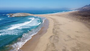 A beach with clear blue water, a sandy shore, and rocky outcrops. - 3 bedroom villa in quiet complex with swimming pools / private wifi / UKTV (Corralejo)