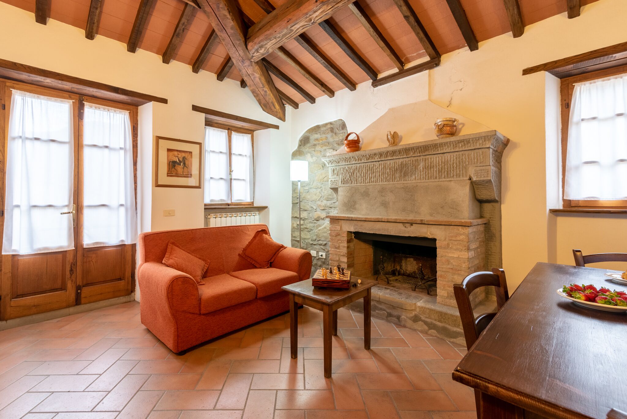 A cozy living room with a fireplace, a sofa, a dining table with a plate of strawberries, and wooden beams on the ceiling.
