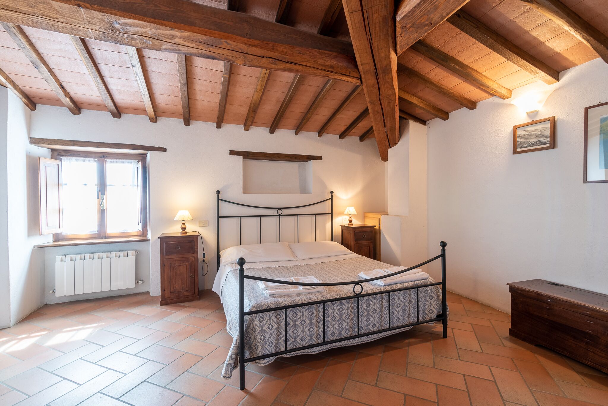 A bedroom with a bed, wooden beams, a window, a radiator, and a bedside table with a lamp.